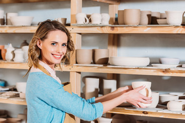 beautiful smiling potter with ceramic dishware on shelves in workshop