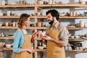 smiling potters talking and holding ceramic dishware in workshop