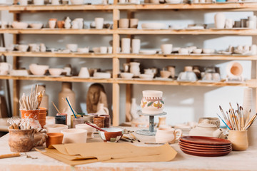 brushes, paints and wooden shelves with ceramics in pottery workshop - obrazy, fototapety, plakaty