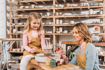 female teacher and child painting ceramic pot in pottery workshop with shelves