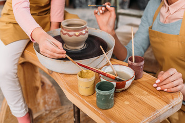 cropped view of teacher and kid painting ceramic pot with brushes in workshop
