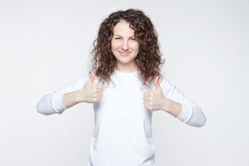 Headshot of happy young Caucasian woman with curly hair in white T-shirt, gesturing thumbs-up, telling that you are doing good job, looking, smiling gently at camera with cheerful confident expression