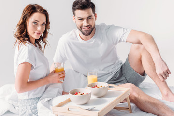 young couple with breakfast on wooden tray looking at camera in bed isolated on grey