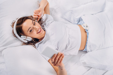 overhead view of young smiling woman in headphones using smartphone in bed