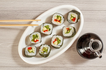 Fototapeta na wymiar Sushi rolls with rice, pieces of avocado, cucumber, red bell pepper and lettuce leaves on ceramic plate, glass bottle with soy sauce