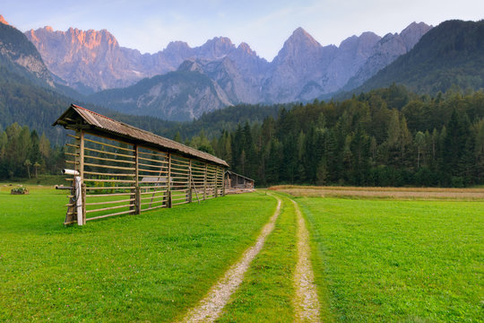 Alpine landscape with dirt road crossing fresh green meadow and traditional hayrack. Dramatic rocky mountain peaks on background. Beautiful nature of Slovenia
