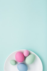 Group of colorful Easter eggs on white plate on turquoise background. Top view, minimal styled greeting card.