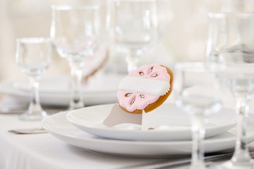 Little glazed gingerbread cookie decorated with pink pattern and white pearls stands on a white plate. There are wine glasses on the background. A festive table is covered with snowwhite tablecloth.