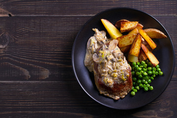 Beef steak diane with mushroom and leek cream sauce, potato fries and green peas in black plate on wooden background. overhead