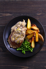Beef steak diane with mushroom and leek cream sauce, potato fries and green peas in black plate on wooden background. overhead
