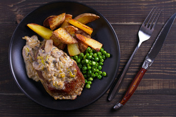 Beef steak diane with mushroom and leek cream sauce, potato fries and green peas in black plate on wooden background. overhead