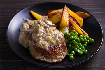 Beef steak diane with mushroom and leek cream sauce, potato fries and green peas in black plate on wooden background