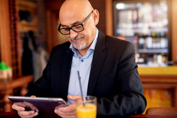 Mature professional focused businessman is using a tablet and drinking juice in the coffee shop.