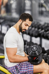 Fototapeta na wymiar Strong sexy bearded man wears white t-shirt and headphones does arm biceps workout with dumbbells in the gym