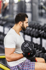 Fototapeta na wymiar Handsome sexy bearded man wears white t-shirt and headphones does arm biceps workout with dumbbells in the gym
