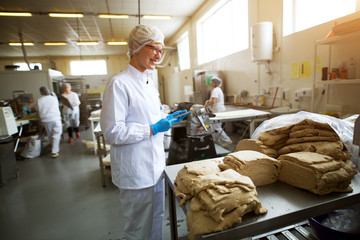 Young cheerful female worker in sterile cloths holding a tablet and checking the quality of cookie dough mixture before further processing.