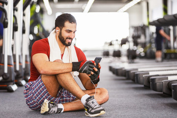 Fototapeta na wymiar Bearded muscular man wears red t-shirt sitting on floor while rest with towel on neck, using smartphone in the gym