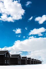 the same dark houses stand in a row, in winter on a sunny day. The sky is blue with clouds. Snow covered earth. Minimalism.