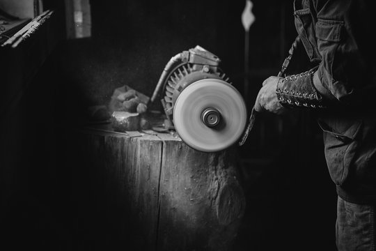 The worker grinds a metal detail on the grinder with an abrasive wheel. The old grinder machine is fixed on the tree trunk saw cut - Powered by Adobe
