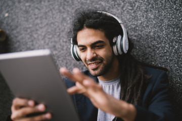 Close up picture of young stylish man with dreadlocks using tablet and listening to music on his headphones while smiling.