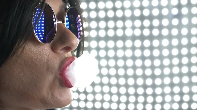 Hot brunette in round sunglasses, chewing gum and blowing a bubble. 4k, slow motion.