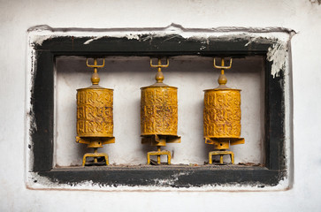Three metal prayer wheels of yellow color, with mantras "om mani peme hung" in a wooden frame of black color on a white wall of the Buddhist temple.