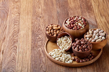 Mixed nuts in brown bowls on wooden tray over rustic background, close-up, top view, selective focus.