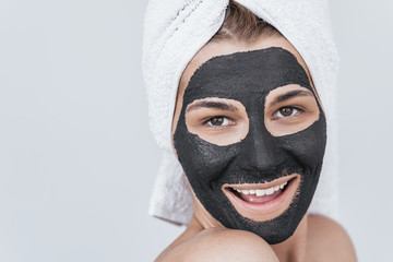 Closeup shot of beautiful smiling young female doing beauty treatment on her face, clean skin with black organic clay, looks at the camera with toothy healthy smile. Woman applying organic facial mask