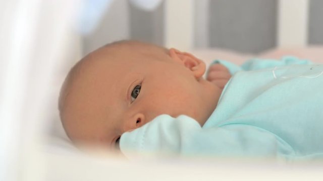 Close-up shot of quiet sleepy newborn baby in blue onesie lying in bed, crib mobile spinning overhead