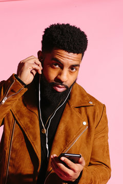 Cool n with beard holding his cell phone and  touching headphone in ear, isolated on pink studio background