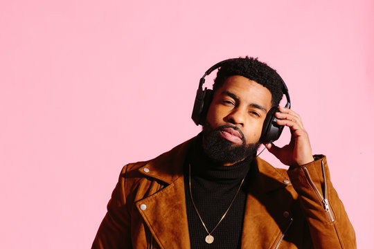 Cool African American man with beard holding his headphones enjoying listening to music, isolated on pink studio background