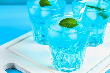 Blue cocktail with ice and mint in glasses on a white wooden board on a blue table.