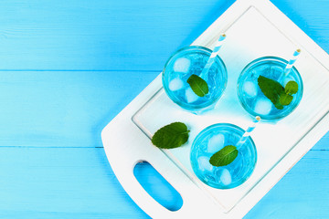 Blue cocktail with ice and mint in glasses on a white wooden board on a blue table. Top view.