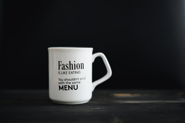 lettering of inspirational quote FASHION IS LIKE EATING, YOU SHOULDN'T STICK WITH THE SAME MENU on white mug, on the wooden black table and black background