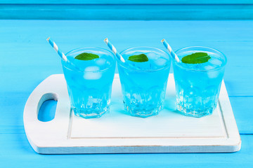 Blue cocktail with ice and mint in glasses on a white wooden board on a blue table.