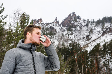 Young man tourist drinking tea from a thermos and smiles on mountains and forest background.