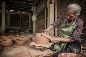 A woman potter in a plaid shirt and green apron beautifully sculpts a deep bowl of brown clay and cuts off excess clay on a potter's wheel