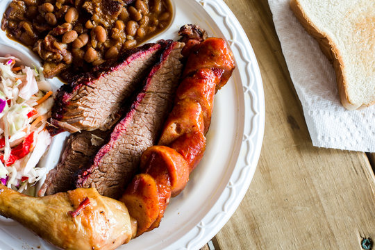 Overhead of a full plate of smoked chicken, sausage and barbecue beef brisket with baked beans and fresh coleslaw on wooden table with white bread on a paper napkin in the background.