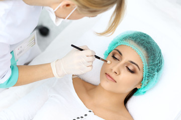 Professional beautician is doing cosmetic procedure at light medical background touching patient's face with brush, closeup.  Cosmetology treatment