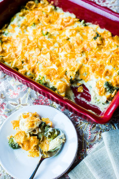 Overhead shot of browned chicken divan casserole in red baking dish on gray floral linen with individual serving on white plate with silver spoon.
