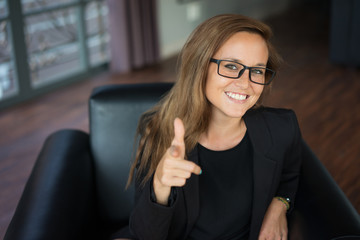 Closeup portrait of smiling young beautiful brown-haired woman looking at camera, pointing finger at viewer and sitting in armchair in hotel lobby. Encouragement concept. Front view.