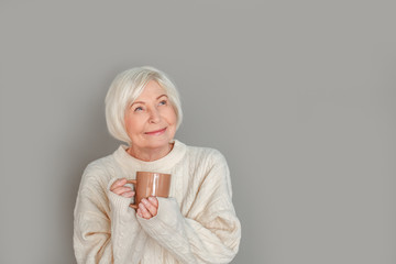 Senior woman in sweater studio isolated on grey wall holding cup with hot milk