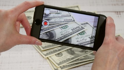 The girl's hands hold a smartphone, with which she removes hundred-dollar bills and a purse lying on the table - 2. Close-up.