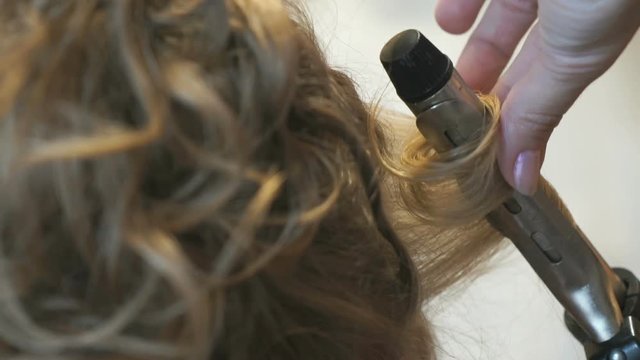 Stylist makes the curls to an attractive brown-haired using a hot curling