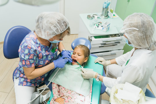 Child with a mother at a dentist's reception. The girl lies in the chair, behind her mother. The doctor works with an assistant. There is an operation to fill the tooth.