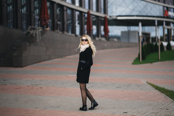 Beautiful young girl with blond wavy hair in a black coat against the backdrop of modern buildings