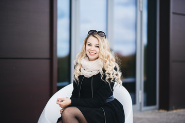 Beautiful young girl with blond wavy hair in a black coat sits on background of modern building