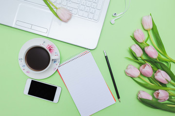 Female blogger workplace with laptop, pink tulips, tea cup, mobile phone, open notebook and black pencil on green background. Top view with empty space for your text, greetings