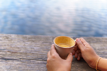 asian female hand  holding  white coffee  or tea cup  on the wooden plank near pond or lake.