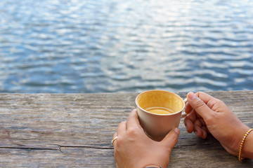 asian female hand  holding  white coffee  or tea cup  on the wooden plank near pond or lake.